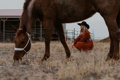 a woman in a red dress and cowboy hat sitting next to a brown horse on dry grass