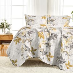 a bed covered in a white and yellow floral quilt next to a table with flowers on it