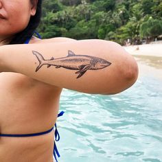 a woman with a fish tattoo on her arm next to the water and trees in the background