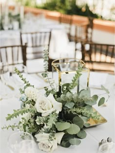 an arrangement of flowers and greenery sits on a table at a wedding reception in the sun