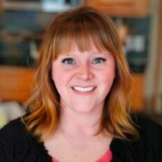 a woman with red hair smiling at the camera in a kitchen area that has wooden cabinets and wood floors