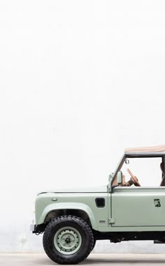 a light green jeep parked in front of a white wall with two people inside it