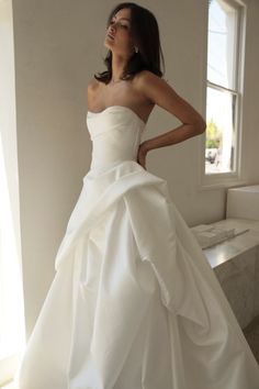 a woman in a white wedding dress standing by a window with her hands on her hips