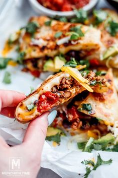 a person holding up a slice of pizza with toppings on it and other food items in the background