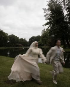 a man and woman dressed in wedding attire running across grass next to a body of water