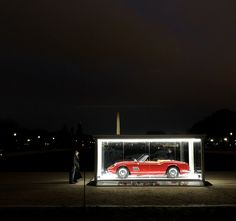 a man standing next to a red car in a display case at night with the lights on