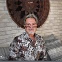 a man wearing a shirt and glasses standing in front of a wall with a clock on it's face