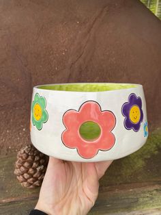 a hand holding up a bowl with flowers painted on the side and a pine cone in the background