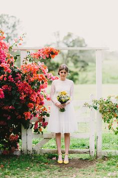 a woman in a white dress standing next to pink and red flowers on a fence