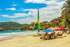 people are sitting under umbrellas on the beach near the water and sailboats in the ocean