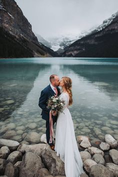 a bride and groom kissing on the shore of a lake surrounded by rocky shores with snow capped mountains in the background