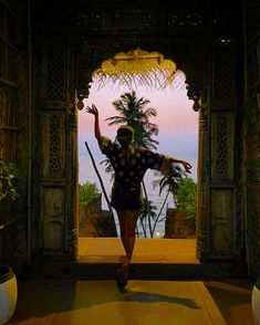 a woman is walking through an archway with her arms in the air and palm trees behind her