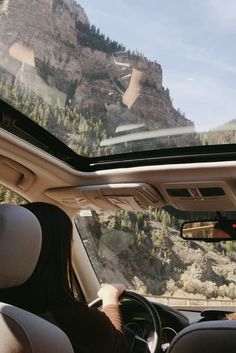 a woman driving a car down a road next to a mountain