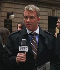 a man in a trench coat and tie holding a microphone while talking to the media