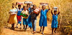 several children carrying boxes on their heads and walking down a dirt path through tall grass