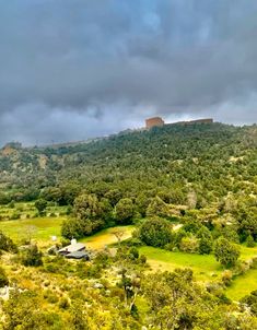 an aerial view of a mountain with trees and grass in the foreground, on a cloudy day