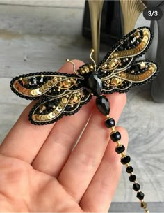 a hand holding a black and gold dragonfly brooch with beads on it's wings