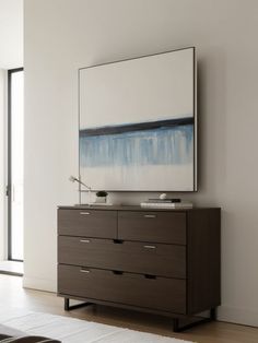 a large painting hangs on the wall next to a dresser in a room with white walls