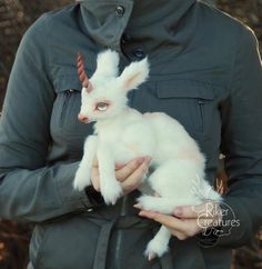 a woman holding a baby white goat in her arms and wearing a jacket with an unicorn horn on it's head