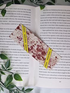 an open book with some blood and yellow tape on top of it next to green leaves