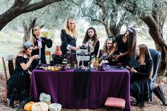 a group of women sitting around a table covered in halloween food and drinks, all dressed up