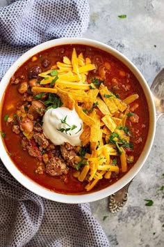 a white bowl filled with chili and cheese on top of a blue towel next to a spoon