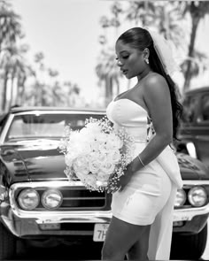 a woman in a white dress holding a bouquet and standing next to a black car