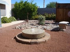 a stone fountain in the middle of a graveled area with trees and bushes behind it