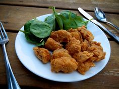 a white plate topped with fried chicken next to spinach leaves and silverware on top of a wooden table