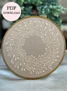 a hand embroidered hoop with white flowers and leaves on it, in front of a potted plant