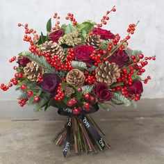 a bouquet of flowers with berries, pine cones and holly on the top is sitting in front of a white wall