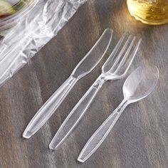 three plastic forks and two spoons on a wooden table next to a jar of pickles