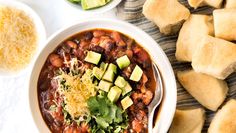 a bowl of chili with avocado, cheese and tortilla bread on the side