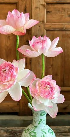 three pink flowers in a green vase on a table next to a wooden wall and door