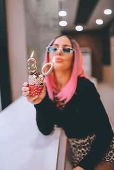 a woman with pink hair holding up a cupcake