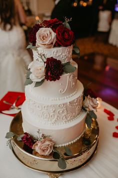 a white wedding cake with red flowers and greenery on the top is sitting on a gold platter