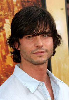 a man with long hair standing in front of a poster and looking at the camera