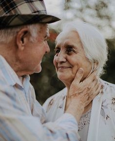 an older man and woman standing next to each other in front of some trees with their faces close together