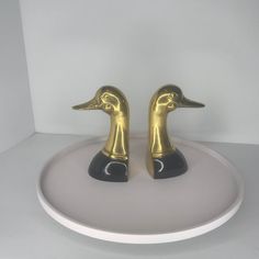 two golden ducks sitting on top of a white plate in front of a white wall