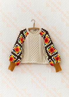 a sweater hanging on a clothes hanger against a white quilted wall with an orange, yellow, and green flower design