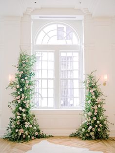 an arch with flowers and greenery in front of a window on a wooden floor