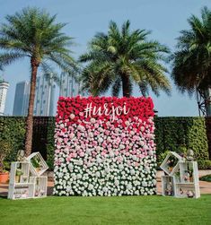 an outdoor wedding setup with flowers on the wall and white chairs in front of palm trees