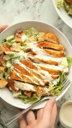 a person is holding a bowl with chicken and lettuce on it, while another hand holds a fork in front of the bowl