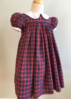 "This homemade dress was very nicely constructed and finished. Made from a poly/cotton blend, it is a woven tartan in red, blue, black, green and white. The waistline is high - an Empire style. The front bodice is finished with a very narrow bias self piping. The skirt below is gathered and mid-calf length. Sleeves are short and very puffed. Cuff is white and finished with self piping. Collar is likewise white with piped edges. It is round and wide. Back bodice sits lower than front and closure Fitted Red Smock Dress, Fitted Classic Plaid Dress With Short Sleeves, Classic Fitted Plaid Dress With Short Sleeves, Fitted Lined Plaid Dress, Plaid Smocked Short Sleeve Dress, Short Sleeve Plaid Smock Dress, Fitted Plaid Dresses With Lining, Classic Short Sleeve Plaid Dress, Classic Fitted Plaid Dress