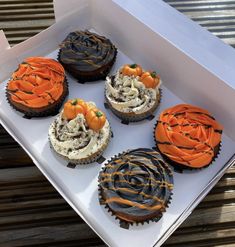 six decorated cupcakes in a white box on a wooden table with brown and orange icing