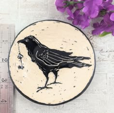 a black bird is sitting on a plate next to some purple flowers and measuring tape