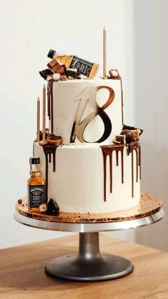 a three tiered white cake with chocolate drips and bottles on the side, sitting on top of a wooden table