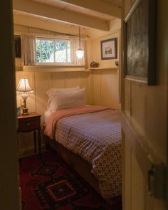 a small bedroom with a bed, nightstand and lamp in it's corner next to a window