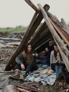 three people sitting on the ground in front of a pile of sticks and wood logs