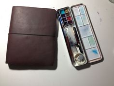 an open brown leather case next to a painting brush and watercolors in it